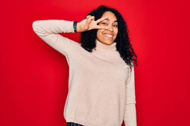 Jeune femme frisée afro-américaine portant un pull à col roulé décontracté sur fond rouge Faire symbole de paix avec les doigts sur le visage, sourire joyeux montrant la victoire
 - Photo, image