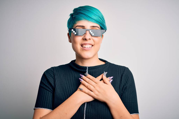 Young woman with blue fashion hair wearing thug life sunglasses over white background smiling with hands on chest with closed eyes and grateful gesture on face. Health concept. - Foto, afbeelding
