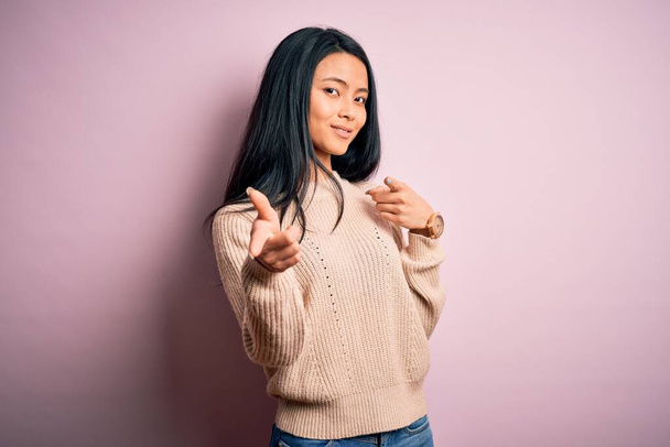 Молодая красивая китайская женщина носит случайный свитер на изолированном розовом фоне указывая пальцами на камеру с счастливым и смешным лицом. Хорошая энергия и вибрации
. - Фото, изображение