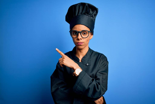 Young african american chef woman wearing cooker uniform and hat over blue background Pointing with hand finger to the side showing advertisement, serious and calm face - Zdjęcie, obraz