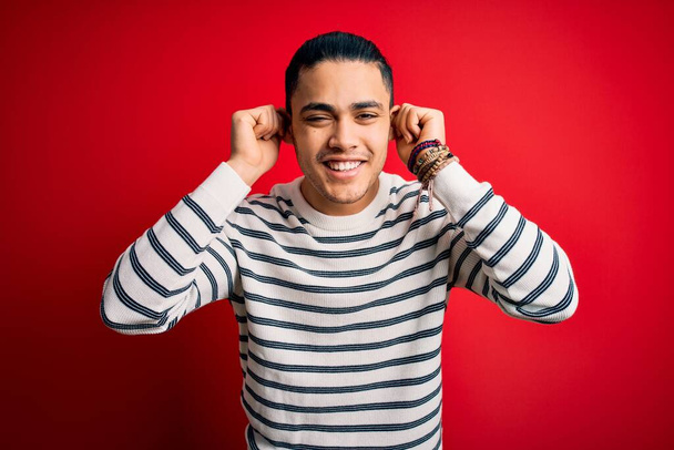 Молодой бразилец в обычной полосатой футболке, стоящей на изолированном красном фоне, улыбаясь, дергая за уши пальцами, смешной жест. Задача прослушивания
 - Фото, изображение