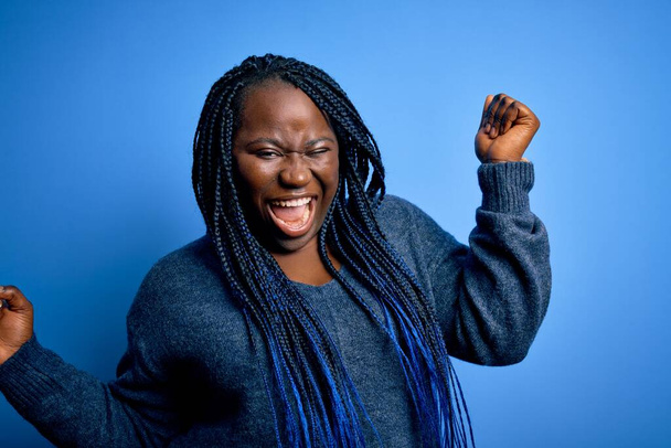 Afroamerykanka plus size kobieta z warkoczami ubrana w luźny sweter na niebieskim tle Taniec szczęśliwy i wesoły, uśmiechnięty poruszający się swobodnie i pewnie słuchając muzyki - Zdjęcie, obraz