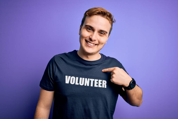 Junge hübsche rothaarige Mann trägt freiwillige T-Shirt über isolierten lila Hintergrund zeigt mit dem Finger auf sich selbst lächelnd glücklich und stolz - Foto, Bild