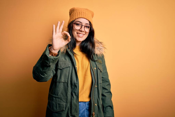 若いブルネットの女性は、黄色の孤立した背景に帽子をかぶった眼鏡と冬のコートを身に着けている肯定的な笑顔手と指でokサインをします。成功した表現. - 写真・画像