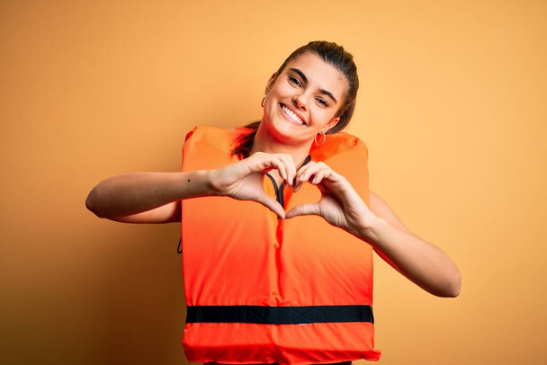 Mladá krásná brunetka žena na sobě oranžové bezpečné záchranné vesty přes žluté pozadí s úsměvem v lásce dělá srdce symbol tvar s rukama. Romantický koncept. - Fotografie, Obrázek