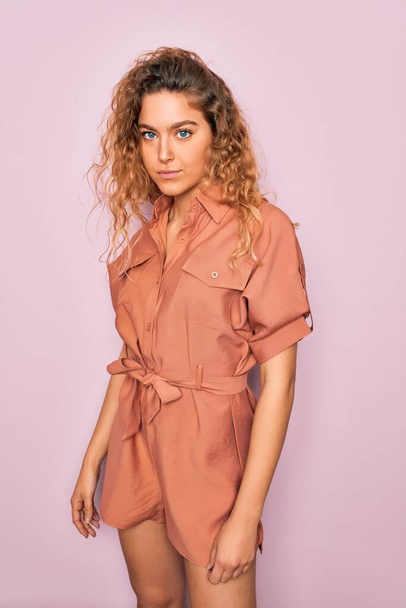 Young beautiful blonde woman with curly hair wearing casual dress standing over isolated pink background - Photo, Image