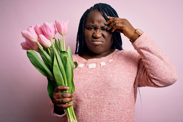 Młoda Afroamerykanka plus size kobieta z warkoczami trzymająca bukiet różowych tulipanów kwiat zaniepokojony i zestresowany o problem z ręką na czole, nerwowy i zaniepokojony kryzysem - Zdjęcie, obraz