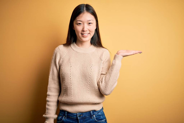 Joven hermosa mujer asiática vistiendo suéter casual sobre amarillo aislado fondo sonriente alegre presentando y señalando con la palma de la mano mirando a la cámara
. - Foto, Imagen