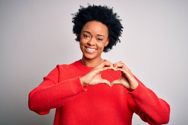 Junge schöne afroamerikanische Afro-Frau mit lockigem Haar, die einen roten lässigen Pullover trägt und verliebt lächelt und Herzsymbolform mit den Händen macht. Romantisches Konzept. - Foto, Bild