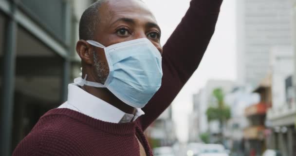 African American man out and about in the city streets during the day, wearing a face mask against air pollution and covid19 coronavirus, rising his hand and hailing a taxi. - Záběry, video