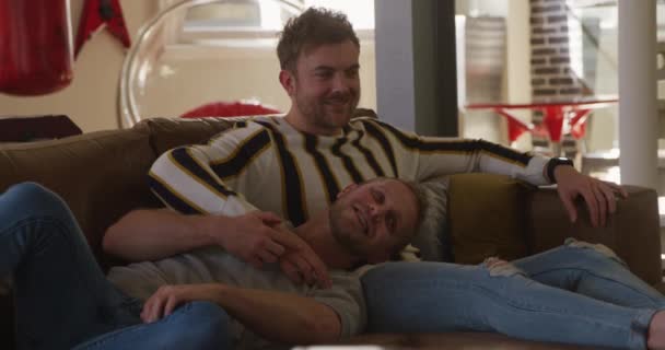 Happy Caucasian male same sex couple relaxing at home together in their living room, sitting on the sofa, embracing and watching TV, one man lying with his head in the lap of the other - Záběry, video