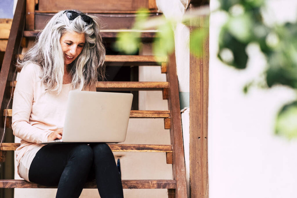 Onnellinen trendikäs valkoihoinen vanhempi nainen käyttää online-tietokonetta internet-yhteyden työskennellä puutarhassa kotona kuin vaihtoehtoinen toimisto älykäs työ teknologia ihmiset
 - Valokuva, kuva