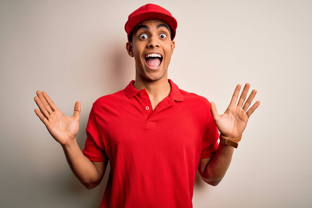 Jovem homem afro-americano bonito vestindo pólo casual e boné sobre fundo vermelho celebrando louco e espantado pelo sucesso com os braços levantados e olhos abertos gritando animado. Conceito de vencedor
 - Foto, Imagem