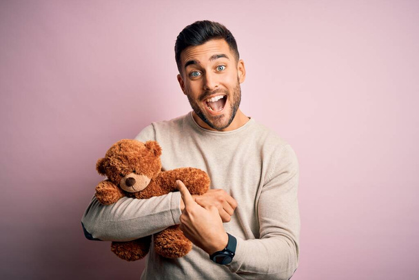 Young handsome man holding teddy bear standing over isolated pink background very happy pointing with hand and finger - 写真・画像