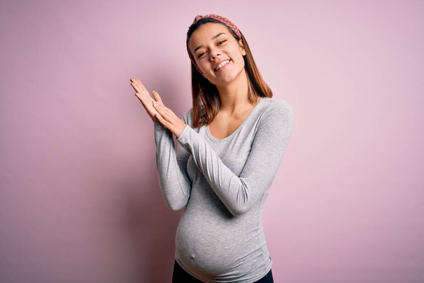 Fiatal gyönyörű tinédzser lány terhes vár baba felett elszigetelt rózsaszín háttér tapsol és tapsol boldog és örömteli, mosolygós büszke kezét együtt - Fotó, kép