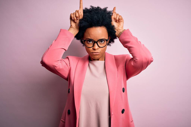 Jovem bela afro-americana afro empresária com cabelo encaracolado usando jaqueta rosa fazendo gesto engraçado com o dedo sobre a cabeça como chifres de touro
 - Foto, Imagem