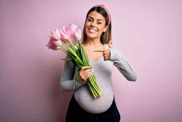 Joven hermosa morena embarazada esperando bebé sosteniendo ramo de tulipanes muy feliz señalando con la mano y el dedo
 - Foto, Imagen
