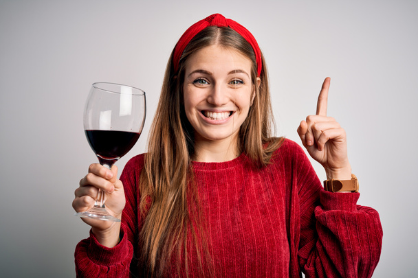 Молодая красивая рыжая женщина пьет бокал красного вина на изолированном белом фоне, удивленный идеей или вопросом указывая пальцем со счастливым лицом, номер один
 - Фото, изображение