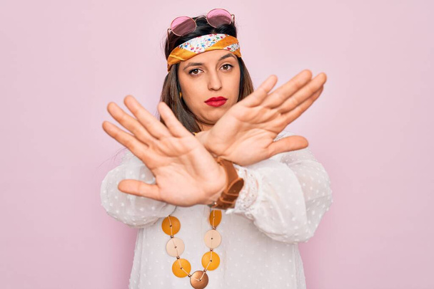 Молода іспаномовна хіпі жінка в стилі моди бухо і сонцезахисні окуляри на рожевому фоні Вираз відторгнення схрещування рук і долонь робить негативний знак, сердите обличчя
 - Фото, зображення