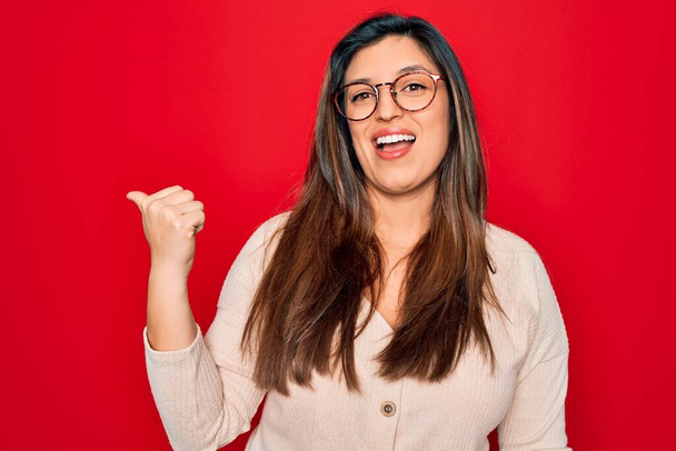 jonge hispanic slimme vrouw dragen bril staan over rood geïsoleerde achtergrond glimlachen met gelukkig gezicht kijken en wijzen naar de zijkant met duim omhoog. - Foto, afbeelding