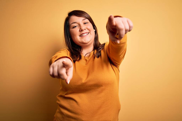 Beautiful brunette plus size woman wearing casual sweater over isolated yellow background pointing to you and the camera with fingers, smiling positive and cheerful - Photo, Image