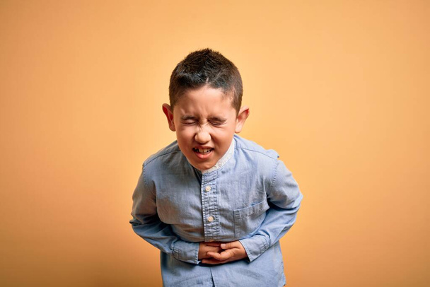 Jeune garçon enfant portant une chemise élégante debout sur fond jaune isolé avec la main sur le ventre parce que les nausées, la maladie douloureuse se sentent mal. Concept Ache
. - Photo, image
