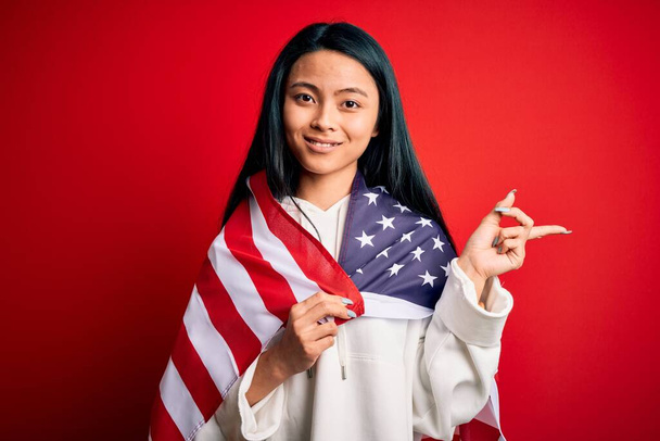 Young beautiful chinese woman wearing United States flag over isolated red background very happy pointing with hand and finger to the side - Foto, afbeelding