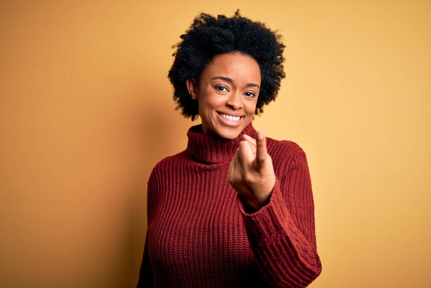 Young beautiful African American afro woman with curly hair wearing casual turtleneck sweater Beckoning come here gesture with hand inviting welcoming happy and smiling - Fotografie, Obrázek