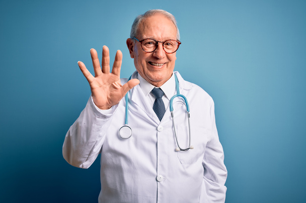Vanhempi harmaahiuksinen lääkäri, jolla on stetoskooppi ja lääketieteellinen takki sinisellä taustalla, osoittaa sormella numero viisi hymyillen itsevarmana ja onnellisena.
. - Valokuva, kuva