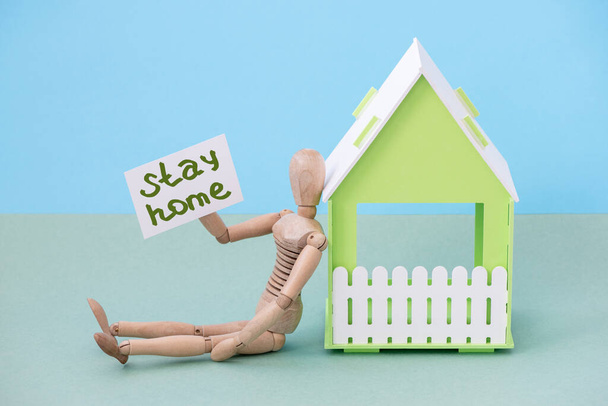 Boneca de boneco de madeira e casa com a inscrição ficar em casa em uma pandemia de coronavírus no fundo de papel
 - Foto, Imagem