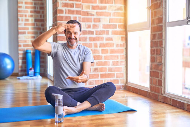 Średniego wieku przystojny sportowiec siedzi na macie robi rozciąganie ćwiczenia jogi na siłowni gestykulując rękami pokazując duży i duży znak rozmiar, symbol pomiaru. Uśmiechnięty patrząc w kamerę. Koncepcja pomiaru. - Zdjęcie, obraz
