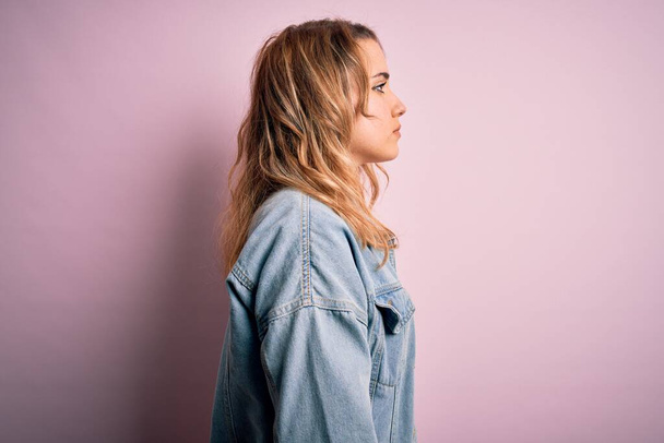 Młoda piękna blondynka nosi luźną kurtkę jeansową stojącą nad różowym tłem patrząc na bok, relaks poza profilem z naturalną twarzą z pewnym uśmiechem. - Zdjęcie, obraz