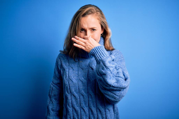 Νεαρή όμορφη ξανθιά γυναίκα που φοράει ζιβάγκο πουλόβερ πάνω από μπλε φόντο μυρίζοντας κάτι βρωμερό και αηδιαστικό, ανυπόφορη μυρωδιά, κρατώντας την αναπνοή με τα δάχτυλα στη μύτη. Άσχημη μυρωδιά. - Φωτογραφία, εικόνα