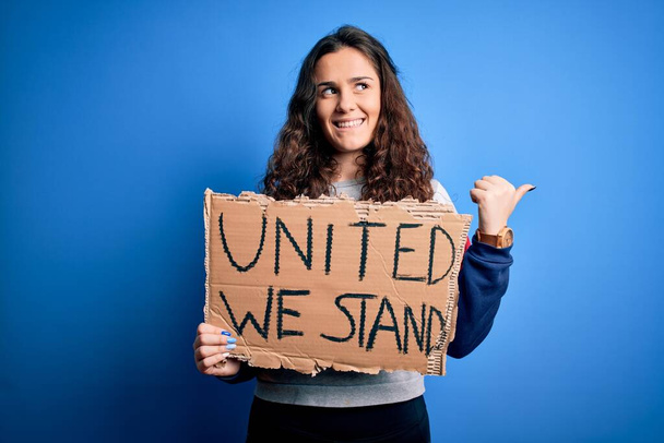 Bella donna attivista che tiene lo striscione con un messaggio in piedi unito su sfondo blu che punta e mostra con il pollice verso l'alto sul lato con il viso felice che sorride
 - Foto, immagini