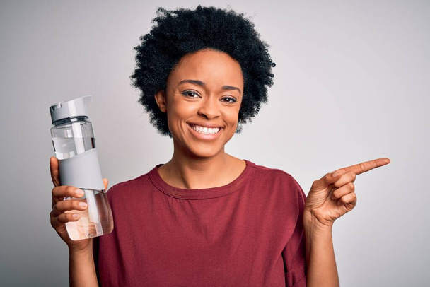 Joven afroamericana afro mujer con pelo rizado bebiendo botella de agua para refrescarse muy feliz señalando con la mano y el dedo a un lado
 - Foto, imagen