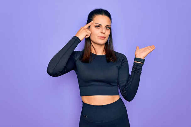 Young beautiful brunette sporty woman wearing casual sportswear over purple background confused and annoyed with open palm showing copy space and pointing finger to forehead. Think about it. - Photo, Image