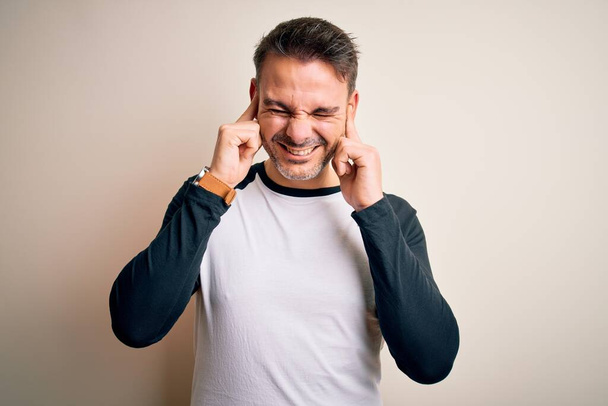 Young handsome man wearing casual t-shirt standing over isolated white background covering ears with fingers with annoyed expression for the noise of loud music. Deaf concept. - Photo, Image