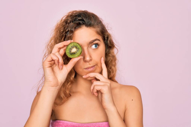 Beautiful woman with blue eyes wearing towel shower after bath holding kiwi fruit over eye serious face thinking about question, very confused idea - Photo, Image