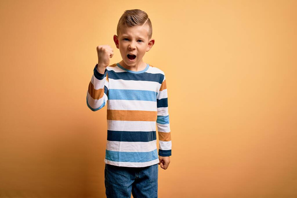 Jonge kleine blanke jongen met blauwe ogen dragen kleurrijke gestreepte shirt over gele achtergrond boos en gek opvoeden vuist gefrustreerd en woedend terwijl schreeuwen van woede. Woede en agressief concept. - Foto, afbeelding