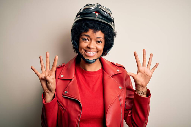 Junge afroamerikanische Afro-Motorradfahrerin mit lockigem Haar trägt Motorradhelm und zeigt mit Finger Nummer neun nach oben, während sie selbstbewusst und glücklich lächelt. - Foto, Bild