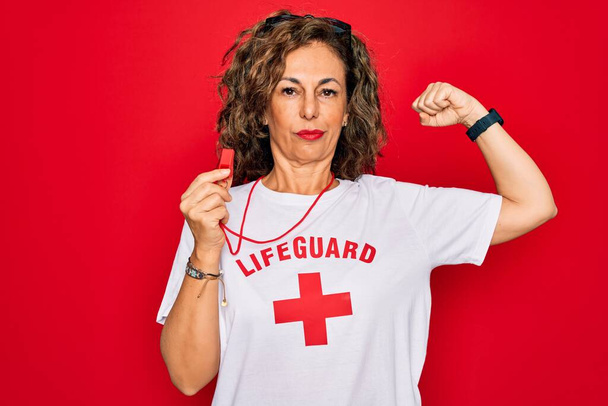 Mujer socorrista de verano de mediana edad que sostiene el silbato sobre el fondo rojo Persona fuerte que muestra el músculo del brazo, confiado y orgulloso del poder
 - Foto, imagen