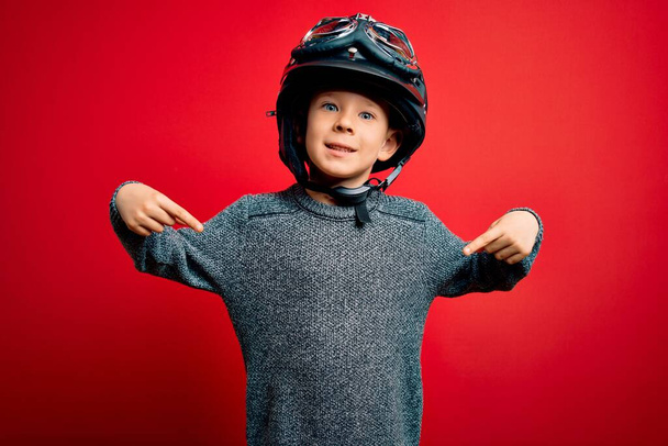 jong klein Kaukasisch kind dragen vintage biker motorfiets helm en googles over rode achtergrond op zoek zelfverzekerd met glimlach op het gezicht, wijzend zich met vingers trots en gelukkig. - Foto, afbeelding
