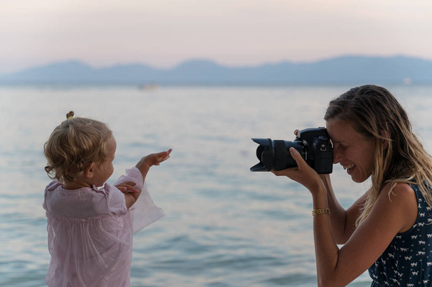 Onnellinen hymyilevä nuori äiti ottaa kuvan hänen pieni lapsi tytär heittää kiviä mereen hämärässä
. - Valokuva, kuva