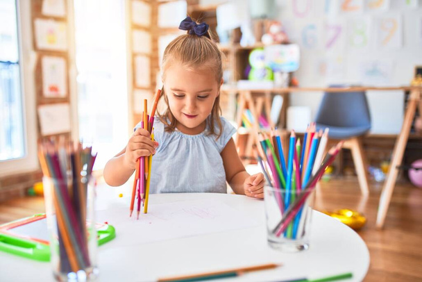 Молодая красивая блондинка девочка наслаждается игрой школы с игрушками в детском саду, улыбаясь счастливая картина с карандашом цвета дома
 - Фото, изображение