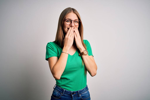Jovem mulher ruiva bonita vestindo casual t-shirt verde e óculos sobre fundo branco rindo e riso embaraçado cobrindo boca com as mãos, fofocas e conceito escândalo
 - Foto, Imagem