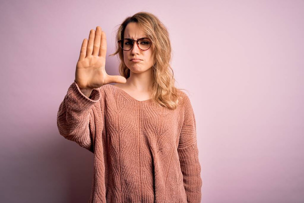 Молодая красивая блондинка носит случайный свитер и очки на розовом фоне делать прекратить петь с ладонью руки. Предупреждающее выражение с негативным и серьезным жестом на лице. - Фото, изображение