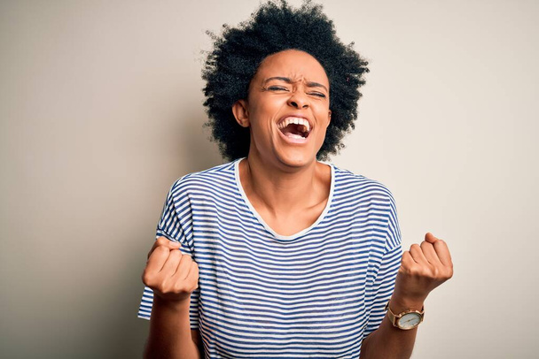 Young beautiful African American afro woman with curly hair wearing striped t-shirt very happy and excited doing winner gesture with arms raised, smiling and screaming for success. Celebration concept. - Foto, afbeelding