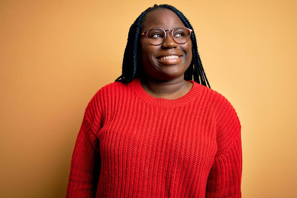 Africký Američan plus velikost žena s copánky na sobě ležérní svetr přes žluté pozadí dívá stranou s úsměvem na tváři, přirozený výraz. Smějící se sebevědomí. - Fotografie, Obrázek