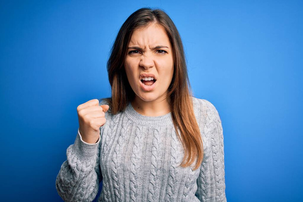 Beautiful young woman wearing casual wool sweater standing over blue isolated background angry and mad raising fist frustrated and furious while shouting with anger. Rage and aggressive concept. - Фото, изображение