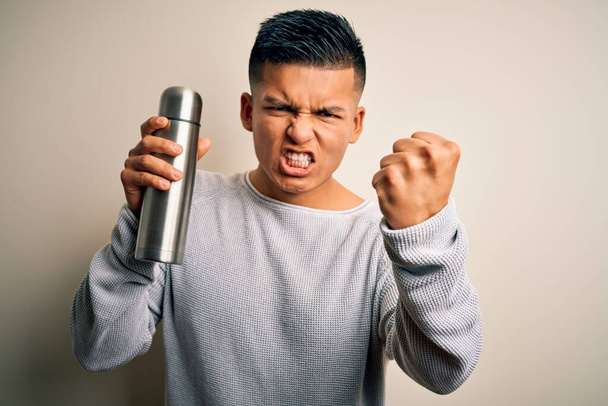 Young handsome latin man holding thermo with water over isolated white background annoyed and frustrated shouting with anger, crazy and yelling with raised hand, anger concept - Fotoğraf, Görsel